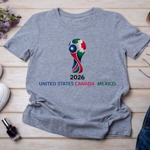 May include: Gray t-shirt featuring a colorful graphic of a soccer ball trophy with the text "2026 UNITED STATES CANADA MEXICO". The design incorporates red, white, blue, and green colors. The shirt is made of a soft material.