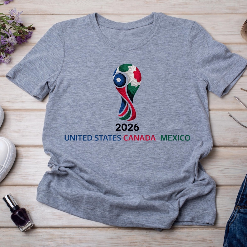 May include: Gray t-shirt featuring a colorful graphic of a soccer ball trophy with the text "2026 UNITED STATES CANADA MEXICO". The design incorporates red, white, blue, and green colors. The shirt is made of a soft material.
