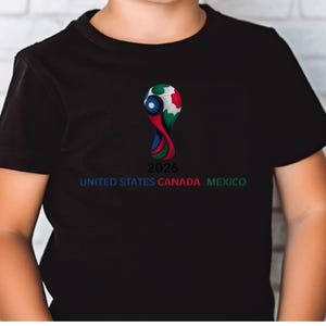 May include: Black t-shirt featuring a colorful graphic of a soccer ball with the text "2026 UNITED STATES CANADA MEXICO" in blue, red, and green. The design is centered on the front of the shirt.
