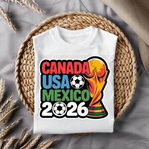 以下が含まれることがあります： 2026 FIFAワールドカップのカラフルなグラフィックがプリントされた白いTシャツ。「CANADA USA MEXICO 2026」の文字が赤、青、緑でデザインされ、サッカーボールとトロフィーが描かれています。