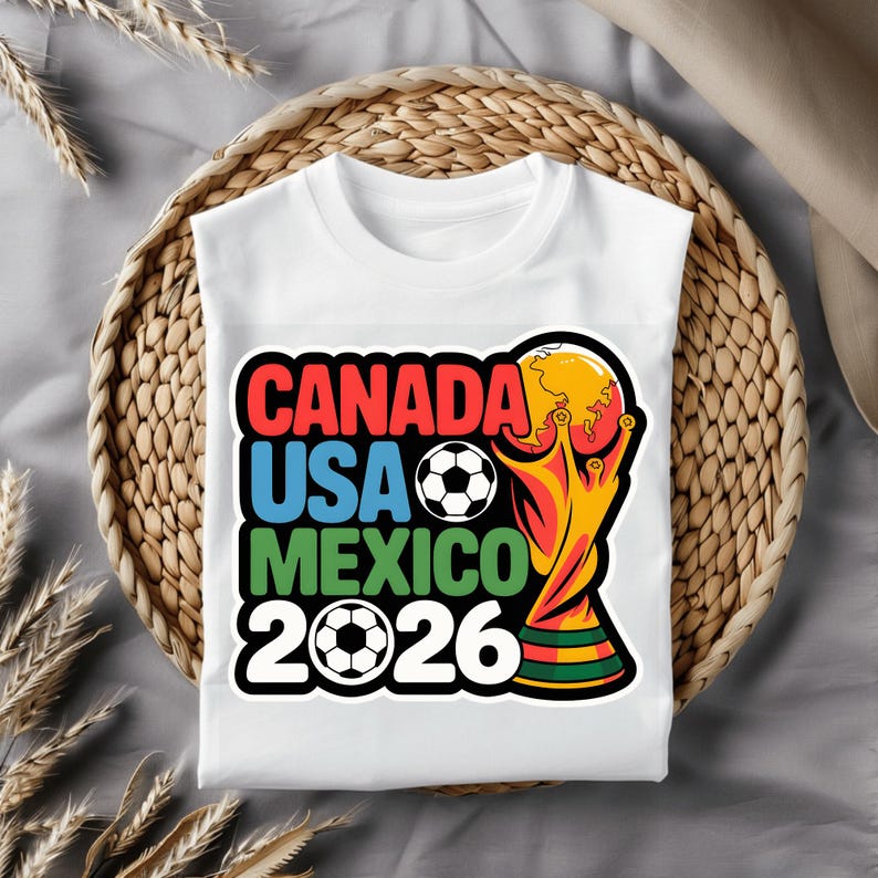 以下が含まれることがあります： 2026 FIFAワールドカップのカラフルなグラフィックがプリントされた白いTシャツ。「CANADA USA MEXICO 2026」の文字が赤、青、緑でデザインされ、サッカーボールとトロフィーが描かれています。