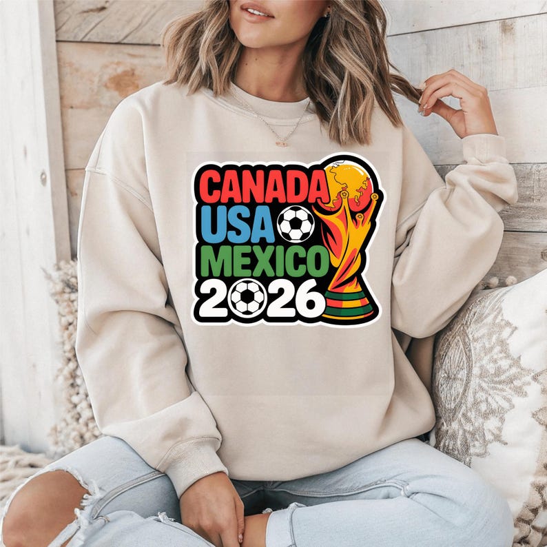 以下が含まれることがあります： 2026 FIFAワールドカップのカラフルなグラフィックがプリントされたベージュのスウェットシャツ。「CANADA USA MEXICO 2026」の文字、サッカーボール、トロフィーのデザインが含まれています。カジュアルなスタイルです。