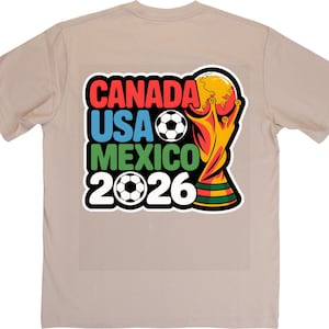 以下が含まれることがあります： 2026 FIFAワールドカップのカラフルなグラフィックが特徴のベージュのTシャツ。「CANADA USA MEXICO 2026」の文字が赤、青、緑、白でデザインされ、サッカーボールとトロフィーが描かれています。