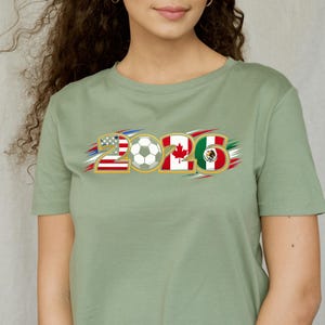 Puede incluir: Camiseta verde salvia con el n&uacute;mero "2026" y un bal&oacute;n de f&uacute;tbol en el centro. Los n&uacute;meros est&aacute;n dise&ntilde;ados con las banderas de Estados Unidos, Canad&aacute; y M&eacute;xico, con detalles en rojo, blanco y azul.