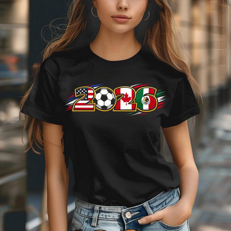 Puede incluir: Camiseta negra con el texto "2026" y un bal&oacute;n de f&uacute;tbol. El dise&ntilde;o incorpora las banderas de Estados Unidos, Canad&aacute; y M&eacute;xico. La camiseta tiene cuello redondo y mangas cortas. Los colores utilizados son rojo, blanco, azul, verde y negro.