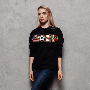 Puede incluir: Sudadera negra con el a&ntilde;o "2026" en un dise&ntilde;o con las banderas de Estados Unidos, Canad&aacute; y M&eacute;xico, junto con un bal&oacute;n de f&uacute;tbol. La persona lleva vaqueros azules.