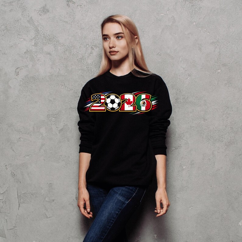 Puede incluir: Sudadera negra con el a&ntilde;o "2026" en un dise&ntilde;o con las banderas de Estados Unidos, Canad&aacute; y M&eacute;xico, junto con un bal&oacute;n de f&uacute;tbol. La persona lleva vaqueros azules.