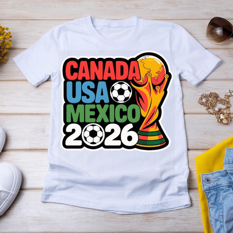 以下が含まれることがあります： 2026 FIFAワールドカップを宣伝するカラフルなグラフィックがプリントされた白いTシャツ。「CANADA」、「USA」、「MEXICO」の文字がそれぞれ赤、青、緑でデザインされ、年号「2026」とサッカーボールが描かれています。
