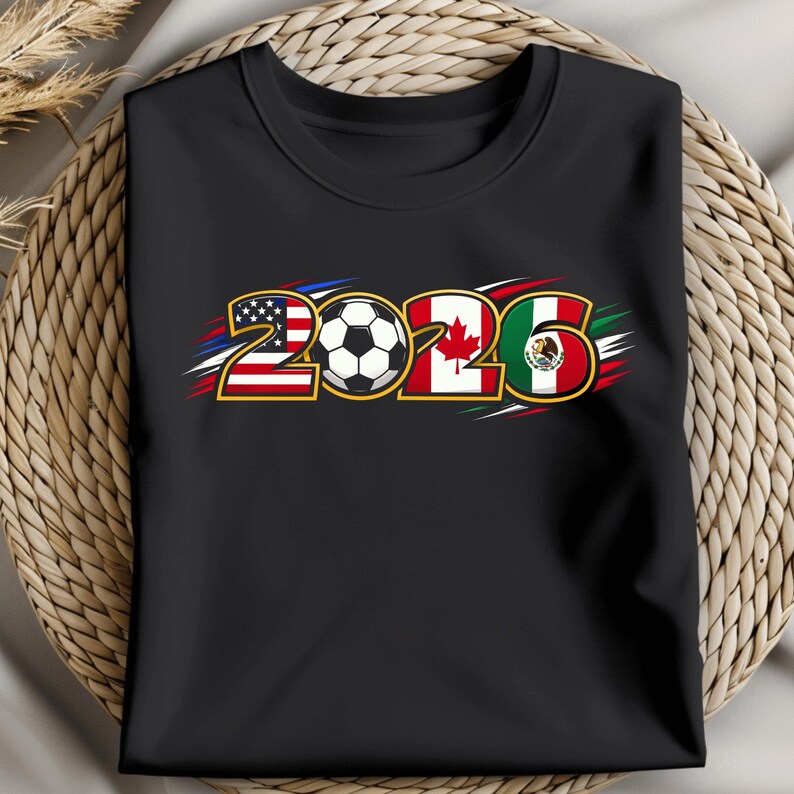 Puede incluir: Camiseta negra con el a&ntilde;o "2026" y un bal&oacute;n de f&uacute;tbol en el centro. Los n&uacute;meros est&aacute;n dise&ntilde;ados con las banderas de Estados Unidos, Canad&aacute; y M&eacute;xico, con detalles en rojo, blanco y azul.