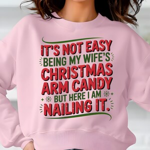 May include: A light pink sweatshirt with the text "IT'S NOT EASY BEING MY WIFE'S CHRISTMAS ARM CANDY BUT HERE I AM NAILING IT" in red and green. The sweatshirt is a crew neck style.