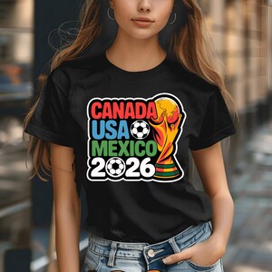 以下が含まれることがあります： 2026 FIFAワールドカップを宣伝するカラフルなグラフィックがプリントされた黒いTシャツ。「CANADA USA MEXICO 2026」の文字、サッカーボール、トロフィーがデザインされています。開催国の国旗も含まれています。