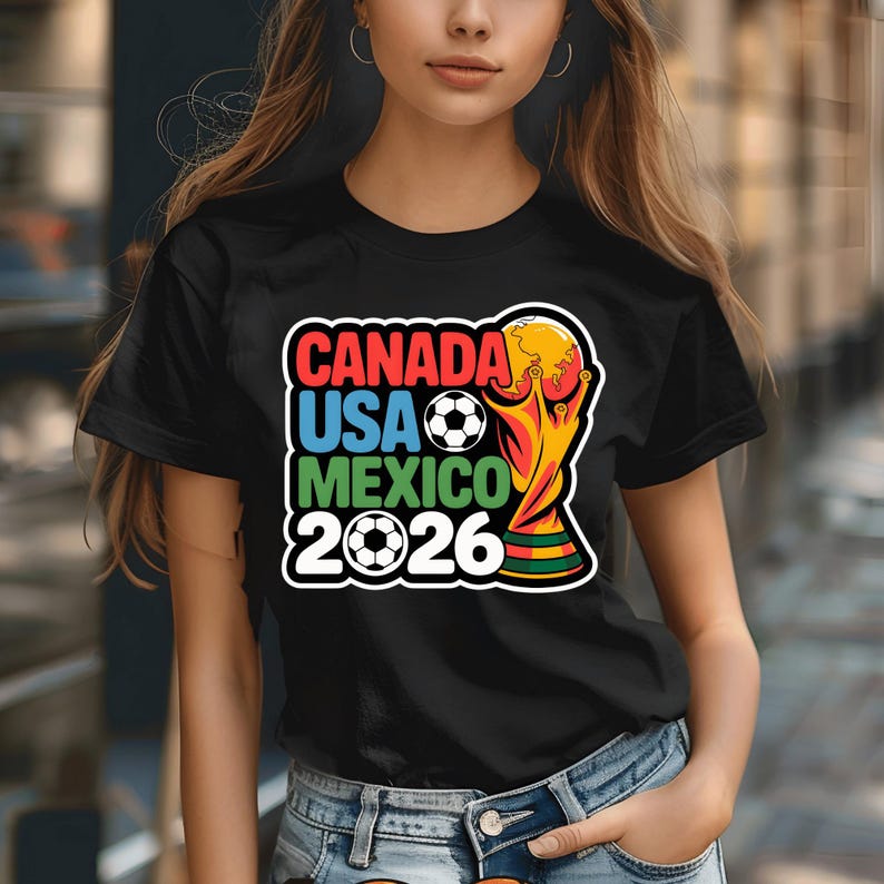 以下が含まれることがあります： 2026 FIFAワールドカップを宣伝するカラフルなグラフィックがプリントされた黒いTシャツ。「CANADA USA MEXICO 2026」の文字、サッカーボール、トロフィーがデザインされています。開催国の国旗も含まれています。