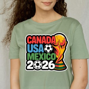以下が含まれることがあります： 2026 FIFAワールドカップのグラフィックがプリントされたセージグリーンのTシャツ。「CANADA USA MEXICO 2026」の文字が赤、青、緑でデザインされ、サッカーボールとトロフィーが描かれています。