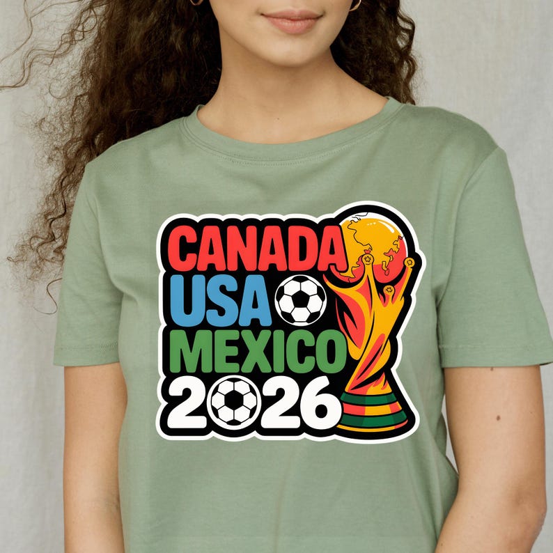 以下が含まれることがあります： 2026 FIFAワールドカップのグラフィックがプリントされたセージグリーンのTシャツ。「CANADA USA MEXICO 2026」の文字が赤、青、緑でデザインされ、サッカーボールとトロフィーが描かれています。