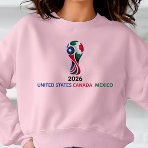 May include: A light pink sweatshirt featuring the 2026 FIFA World Cup logo. The logo is a stylized design in red, green, blue, and white. Below the logo is the number "2026" and the text "UNITED STATES CANADA MEXICO".