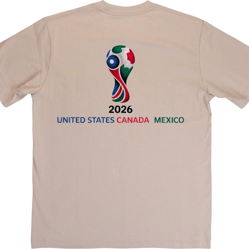 May include: A beige t-shirt featuring a colorful graphic of a soccer ball with the text "2026 UNITED STATES CANADA MEXICO" in blue, green, and red. The shirt is made of a soft material.