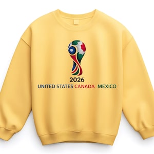 May include: A yellow sweatshirt featuring a graphic of a soccer ball trophy with the text "2026 UNITED STATES CANADA MEXICO". The design is in the center of the sweatshirt.