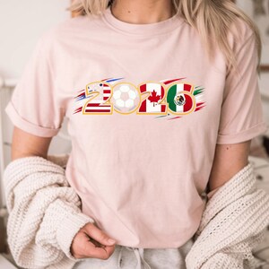 Puede incluir: Camiseta rosa claro con el n&uacute;mero "2026" en contorno dorado. Los n&uacute;meros est&aacute;n dise&ntilde;ados con las banderas de Estados Unidos, Canad&aacute; y M&eacute;xico, con un bal&oacute;n de f&uacute;tbol en el centro. La camiseta es de estilo casual.