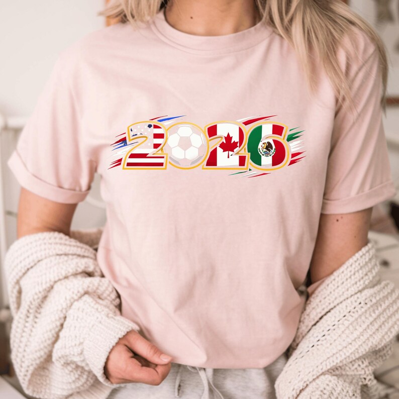 Puede incluir: Camiseta rosa claro con el n&uacute;mero "2026" en contorno dorado. Los n&uacute;meros est&aacute;n dise&ntilde;ados con las banderas de Estados Unidos, Canad&aacute; y M&eacute;xico, con un bal&oacute;n de f&uacute;tbol en el centro. La camiseta es de estilo casual.