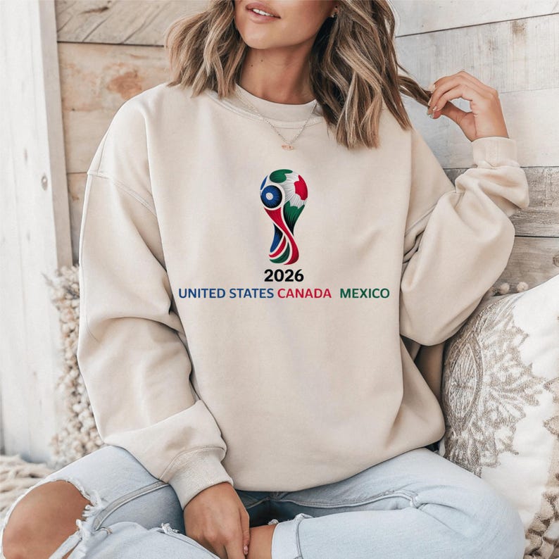 May include: A beige sweatshirt featuring a colorful graphic of a trophy with the text "2026 UNITED STATES CANADA MEXICO". The design is in red, green, and blue. The sweatshirt is a casual style.