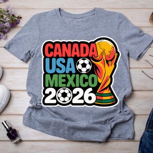 以下が含まれることがあります： 2026 FIFAワールドカップのカラフルなグラフィックがプリントされたグレーのTシャツ。「CANADA USA MEXICO 2026」の文字が赤、青、緑でデザインされ、サッカーボールとトロフィーが描かれています。
