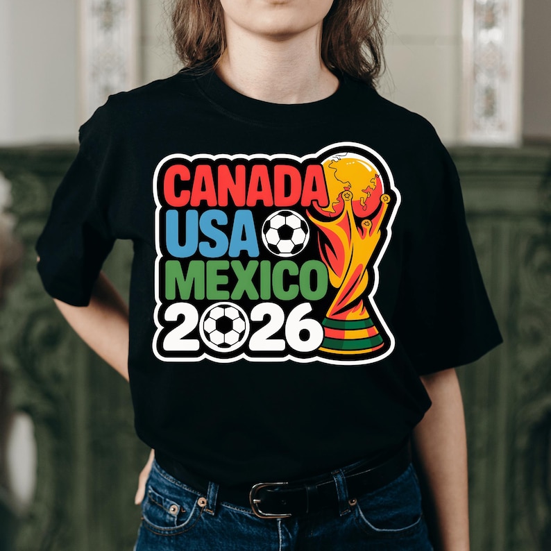 以下が含まれることがあります： 黒いTシャツ。カラフルなグラフィックデザインで、「CANADA」、「USA」、「MEXICO」の文字がそれぞれ赤、青、緑で表示され、サッカーボールと「2026」の年号が描かれています。金色のトロフィーもデザインされています。
