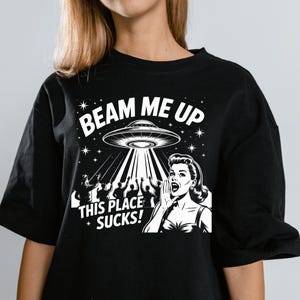 Puede incluir: Camiseta negra con un diseño gráfico retro de un OVNI que teletransporta personas. El texto "BEAM ME UP" y "THIS PLACE SUCKS!" están en blanco. El diseño incluye a una mujer gritando. Un artículo divertido y novedoso.