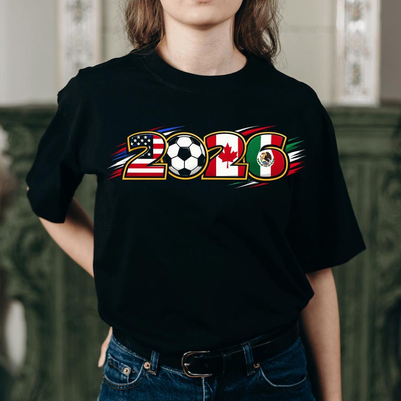 Puede incluir: Camiseta negra con el a&ntilde;o "2026" y un bal&oacute;n de f&uacute;tbol, incorporando las banderas de Estados Unidos, Canad&aacute; y M&eacute;xico. El dise&ntilde;o utiliza colores rojo, blanco y azul.