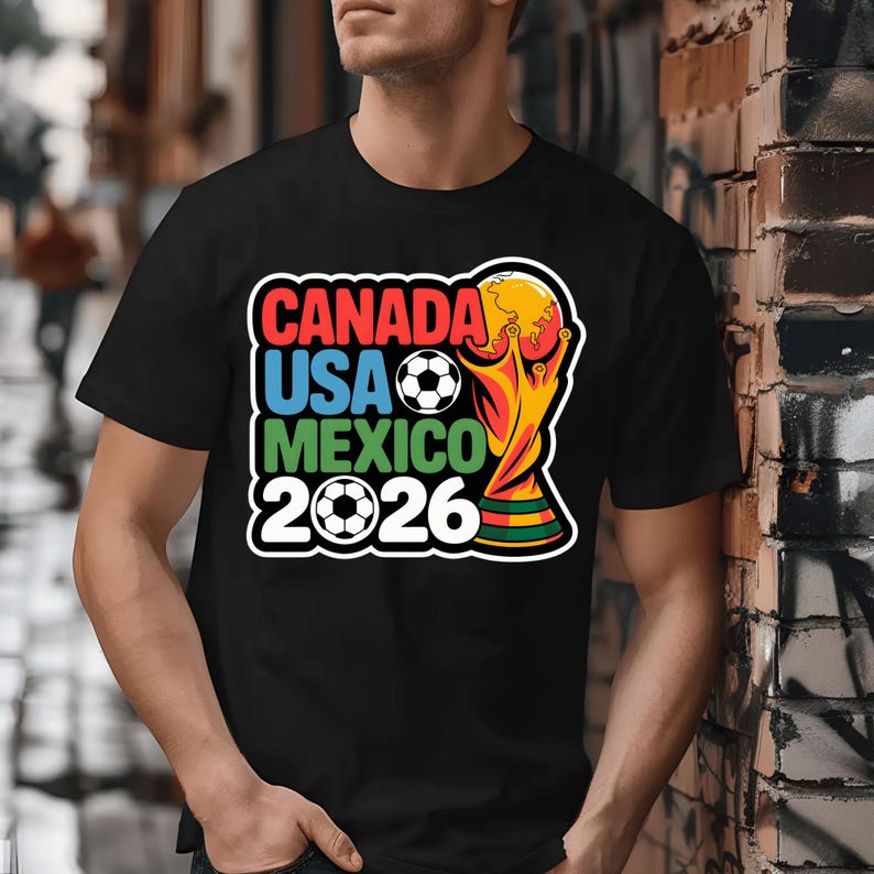 以下が含まれることがあります： 2026 FIFAワールドカップを宣伝するカラフルなグラフィックがプリントされた黒いTシャツ。「CANADA USA MEXICO 2026」の文字が赤、青、緑でデザインされ、サッカーボールとトロフィーが描かれています。