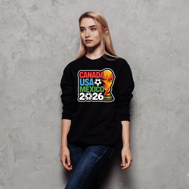 以下が含まれることがあります： 「CANADA USA MEXICO 2026」の文字とサッカーボールとトロフィーのデザインが特徴的な、カラフルなグラフィックが施された黒いスウェットシャツ。青いジーンズを着た人が着用しています。