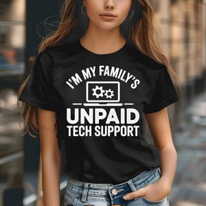 May include: Black t-shirt with the white text "I'M MY FAMILY'S UNPAID TECH SUPPORT". The design includes a laptop icon with gear symbols. The shirt is worn by a person in blue jeans.