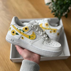 May include: White sneakers with yellow sunflower designs on the side and Nike swoosh. The sneakers are in a white box. The design features yellow sunflowers with green leaves. The sneakers have white laces and a white sole.