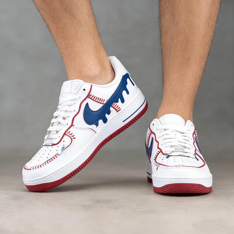 May include: White sneakers with red and blue accents. The shoes feature a blue swoosh design and red stitching, with a red sole. The design incorporates baseball-themed details.