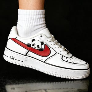 May include: White leather sneaker with a red swoosh and a panda bear graphic. The shoe has black accents and the word "AIR" printed on the side. The sneaker is a low-top style.