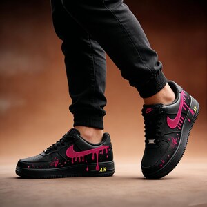 May include: Black sneakers with pink accents and a dripping paint design. The shoes feature a pink Nike swoosh and the word "AIR" in yellow. The sneakers are worn with black pants.