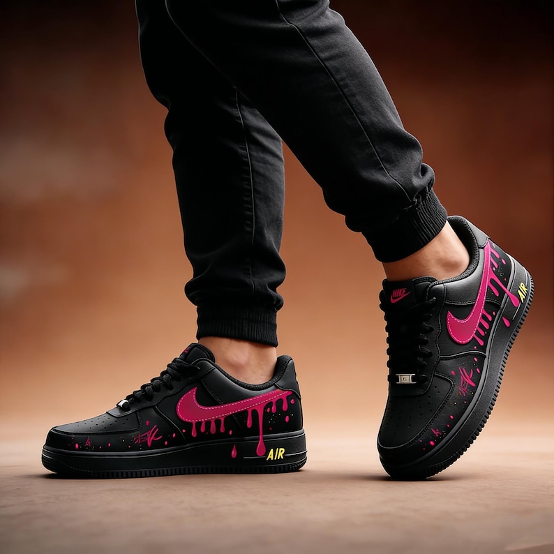 May include: Black sneakers with pink accents and a dripping paint design. The shoes feature a pink Nike swoosh and the word "AIR" in yellow. The sneakers are worn with black pants.