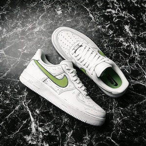 May include: White sneakers with green accents, featuring a unique outlined design. The shoes have a classic low-top style with laces and the iconic swoosh logo in green. The word "AIR" is visible on the side of one shoe.