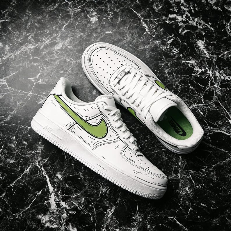 May include: White sneakers with green accents, featuring a unique outlined design. The shoes have a classic low-top style with laces and the iconic swoosh logo in green. The word "AIR" is visible on the side of one shoe.