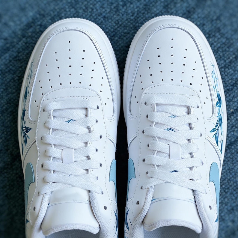 May include: Pair of white sneakers with blue floral accents. The shoes feature a classic design with a perforated toe box and white laces. The blue floral pattern adds a touch of elegance to the athletic footwear.