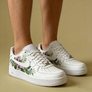 May include: White leather trainers featuring floral patterns in pink, purple, and blue. The trainers have white laces, a white sole, and the Nike swoosh logo. The shoes are a size 9.