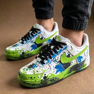 May include: White sneakers with black laces and a green and blue paint splatter design. The shoes have a green Nike swoosh and a green and black sole. The shoes are a low-top style.