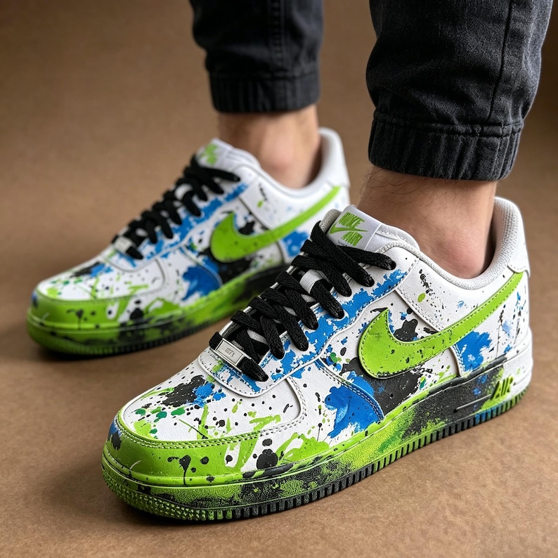 May include: White sneakers with black laces and a green and blue paint splatter design. The shoes have a green Nike swoosh and a green and black sole. The shoes are a low-top style.