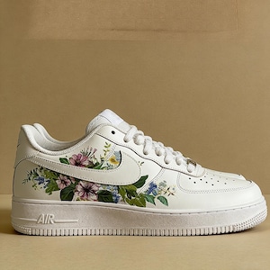 May include: White leather trainer with a floral design. The shoe has a white sole and a colourful floral pattern on the side, including pink, purple, and blue flowers with green leaves. The word "AIR" is visible on the side of the sole.