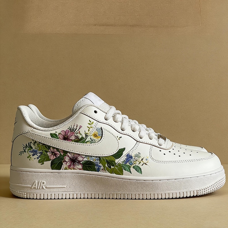 May include: White leather trainer with a floral design. The shoe has a white sole and a colourful floral pattern on the side, including pink, purple, and blue flowers with green leaves. The word "AIR" is visible on the side of the sole.