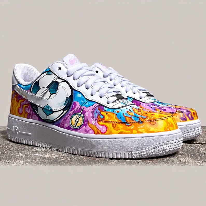 May include: White leather sneakers with a colorful, artistic design. The shoes feature a soccer ball graphic, along with vibrant swirls of blue, yellow, and purple. The soles are white, and the laces are white.