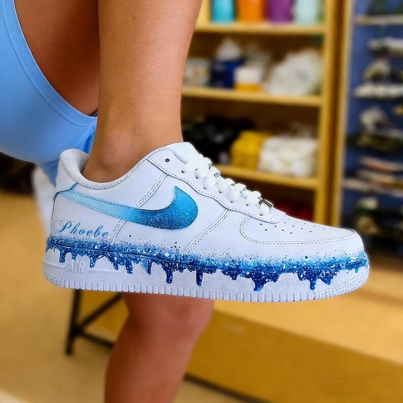 May include: White Nike Air Force 1 sneakers customized with a blue gradient design. The Nike swoosh is blue, and the shoe features a blue drip effect along the sole. The name "Phoebe" is written in cursive on the side.