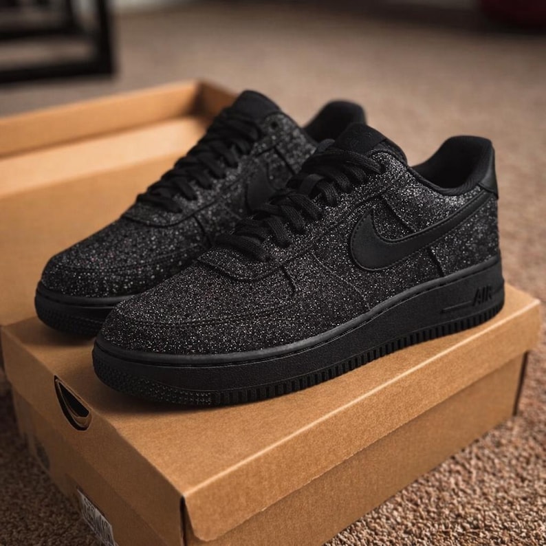 May include: A pair of black sneakers with a glittery, textured finish and black laces. The shoes feature a black Nike swoosh on the side, a solid black sole, and the letters AIR embossed on the heel area. They are resting inside a cardboard box.