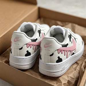 Op de afbeelding: Een paar witte Nike Air Force 1 sneakers met een uniek koe-ge&iuml;nspireerd ontwerp. De schoenen hebben zwarte vlekken, roze accenten en een druipend verfeffect. Het Nike-logo is zichtbaar op de hiel en zijkant.