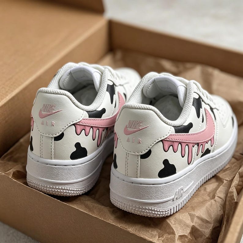 Op de afbeelding: Een paar witte Nike Air Force 1 sneakers met een uniek koe-ge&iuml;nspireerd ontwerp. De schoenen hebben zwarte vlekken, roze accenten en een druipend verfeffect. Het Nike-logo is zichtbaar op de hiel en zijkant.