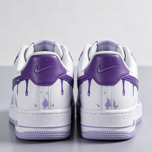 May include: Pair of white and purple sneakers with a low-top design. The sneakers feature a purple heel tab with a Nike logo and purple paint drip details. The soles and accents are also purple.